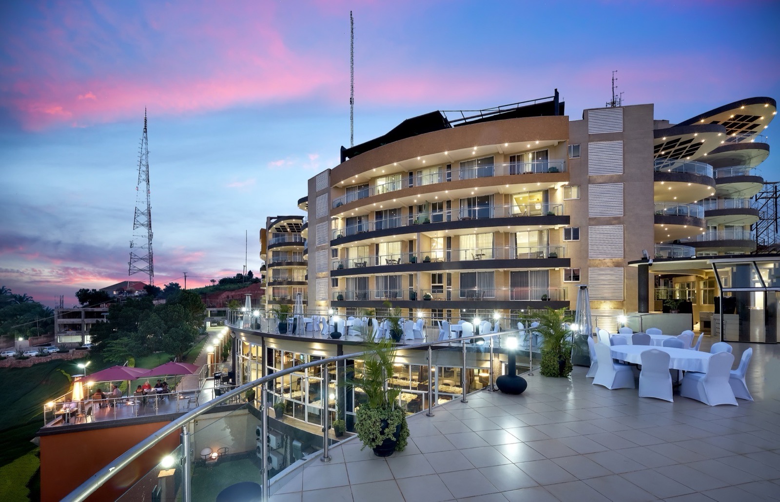 Skyz Hotel Kampala's curved building at dusk, with Skyz Deck rooftop and the ground-floor restaurant terrace lit up