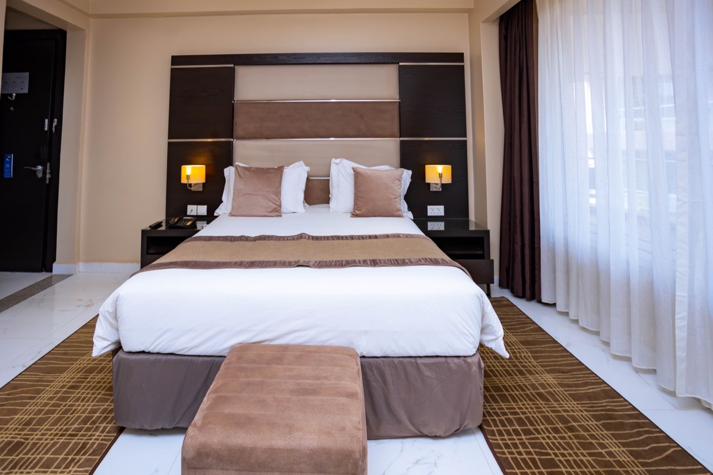 Bed and padded headboard detail in a guest room at Skyz Hotel Kampala, with sheer curtains and soft bedside lighting
