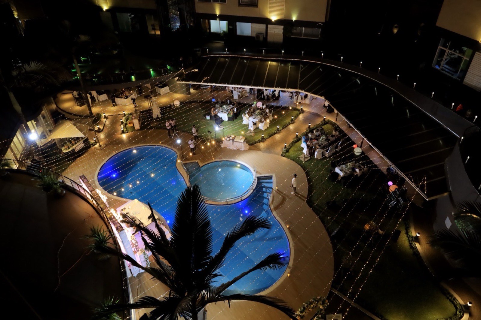 Aerial view of a wedding reception lit with string lights beside the pool courtyard at night, with a live band performing